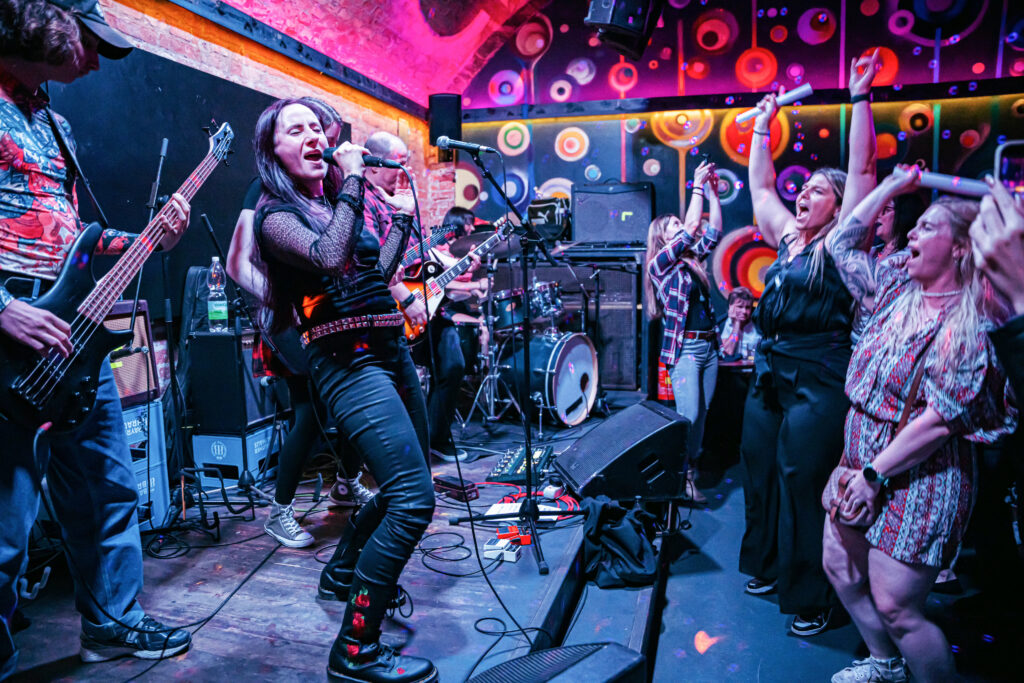School of Rock Schulband beim Live-Auftritt – Sängerin mit Gitarre rockt auf der Bühne mit Band und begeistertem Publikum